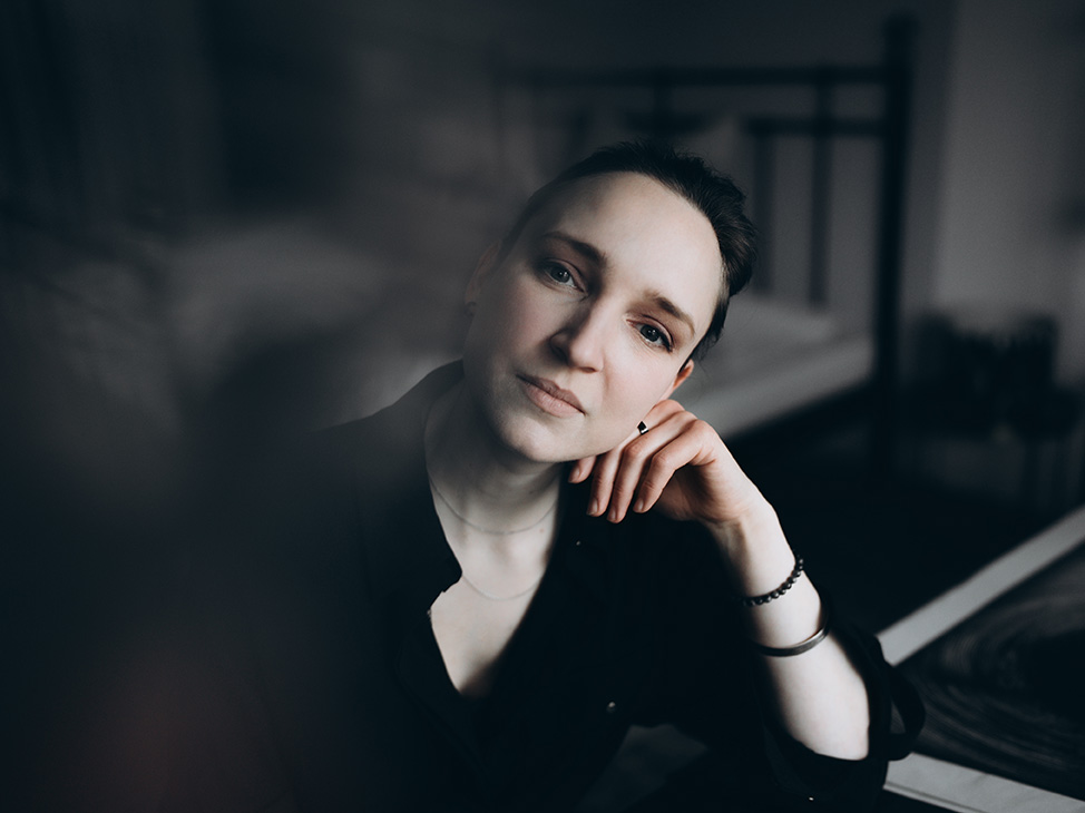 Die Künstlerin mit einem angedeuteten Lächeln und direktem Blick zum/zur Betrachter*in. Den Kopf leicht auf eine Hand gestützt.